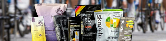 Selection of popular carb and electrolyte drink mixes, including Tailwind, Maurten, Skratch Labs, Precision Hydration, and Thrive EnduraFuel, displayed in front of cyclists during an endurance event.
